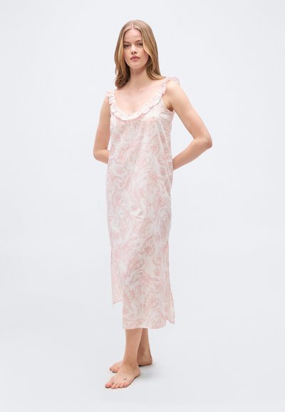 Γυναίκα όρθια ξυπόλητη φορώντας ανοιχτό ροζ ανθισμένο αμάνικο μακρύ φόρεμα με βολάν στον λαιμό και πλευρική σχισμή, μπροστά σε απλό φόντο.