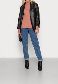 Nainen, joka on pukeutunut mustaan nahkatakkiin, ruosteenväriseen ribattu korkeakaulaiseen villapaitaan, farkkuihin, joissa on käänteet, mustiin nilkkabootseihin, pitäen pientä mustaa käsilaukkua.