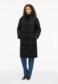 Melnā polsterētā gara mētelis ar augstu apkakli, priekšējo rāvējslēdzēju un sānu kabatām. Materiāls izskatās mīksts ar quilted tekstūru. Modelis valkā zilos džinsus un melnus zābakus.