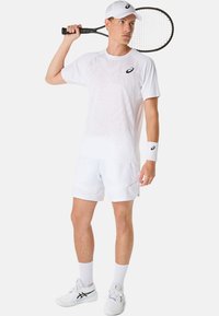 Bílý sportovní outfit skládající se z texturovaného, lehkého trička a odpovídajících šortek, doplněný bílou kšiltovkou a tenisovou raketou.