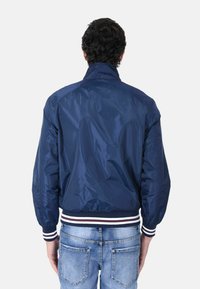 Giubbotto bomber blu navy con colletto alto, vita a coste con strisce rosse e bianche, e materiale liscio e leggero.