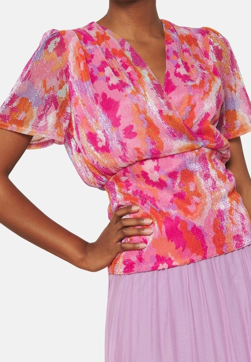 Femme portant un chemisier à motifs rose, orange et violet avec des manches courtes bouffantes et une jupe violet clair.