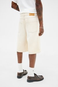 Shorts de mezclilla beige con un corte relajado, que cuentan con bolsillos traseros, un parche de cuero marrón y un dobladillo que cae justo por encima de la rodilla.