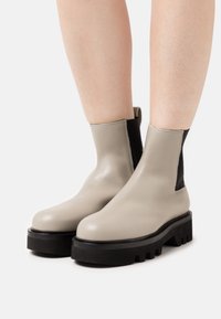 Botas de tobillo de cuero beige con paneles elásticos laterales negros, punta redonda, suela de goma negra gruesa y costuras detalladas a lo largo de las costuras.