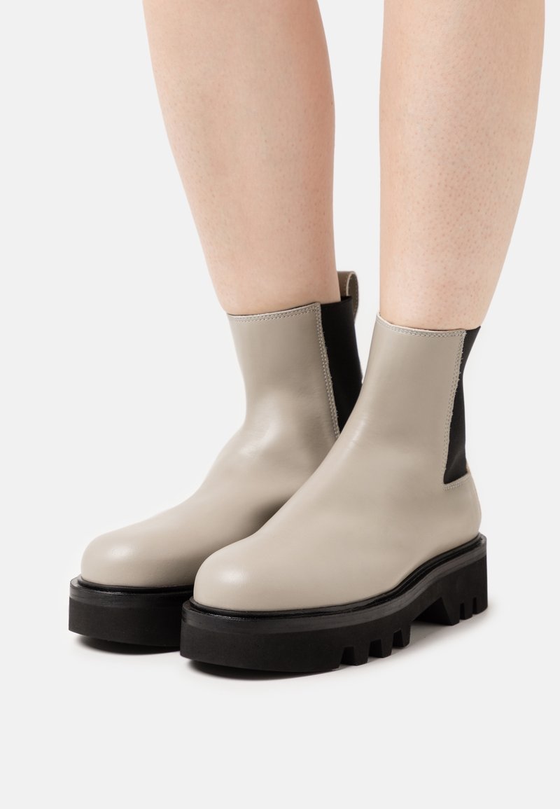 Botas de tobillo de cuero beige con paneles elásticos laterales negros, punta redonda, suela de goma negra gruesa y costuras detalladas a lo largo de las costuras.