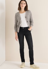 Cárdigan gris texturizado con dos botones, combinado con jeans negros ajustados y zapatillas beige. Camisa blanca debajo del cárdigan. Fondo neutro.