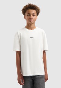 Witte katoenen t-shirt met een ronde hals en korte mouwen, met de tekst "BALLIN'" in zwart over de borst. Draagt een donkere spijkerbroek.