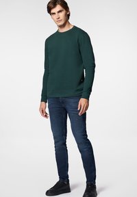 Ciemnozielony sweter z okrągłym dekoltem i długimi rękawami, w połączeniu z wąskimi ciemnymi dżinsami z przetarciami oraz czarnymi sneakersami.
