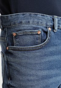 Vaqueros de mezclilla en lavado azul claro, con herrajes de cobre, diseño de cinco bolsillos y sutiles desgastes en la parte frontal.