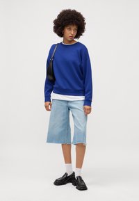 Felpa blu con scollatura tonda e spalle scese, abbinata a pantaloni corti di jeans azzurri, calzini bianchi e scarpe nere.
