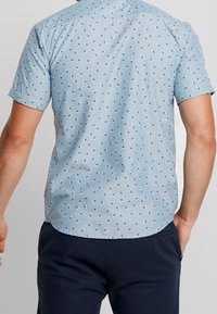 Korte mouwen lichtblauw shirt met een patroon van kleine zwarte en witte driehoeken. Soepele textuur, afgeronde zoom en knoopsluiting bij de kraag.