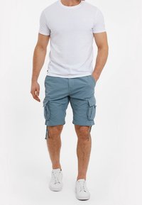 Muž v bílém tričku s krátkým rukávem, modrých cargo šortkách s kapsami a bílých teniskách stojí proti obyčejnému bílému pozadí.