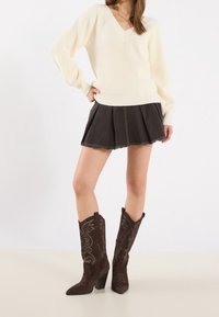 Pull en v crème avec texture côtelée, associé à une jupe plissée en denim noir. Bottes de cowboy en suède marron foncé avec broderies complexes.