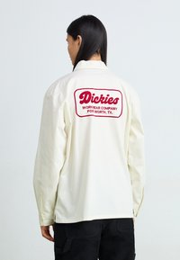 White long-sleeve shirt made of cotton, featuring a red embroidered logo on the back, with buttoned cuffs and a relaxed fit.