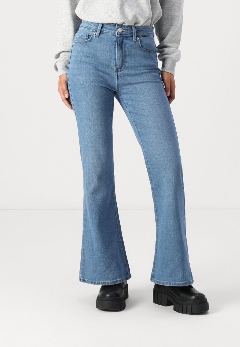 Jean flare taille haute bleu clair portés avec des bottes à plateforme noires et un haut gris à manches longues, vu de mi-cuisse jusqu'aux pieds.
