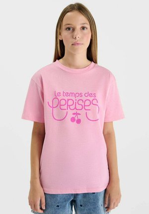 Jeune femme aux longs cheveux portant un t-shirt rose avec le texte français "Le temps des cerises" et un motif de cerise, assorti à un jean bleu.