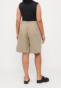 Personne portant un short beige au genou, un haut noir sans manches et des chaussures noires slip-on, debout, dos tourné, sur un fond uni.