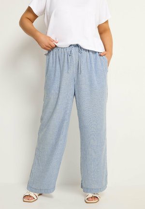 Personne portant un pantalon ample à rayures verticales bleues et blanches avec taille à cordon, t-shirt blanc et sandales blanches ouvertes à l'avant.