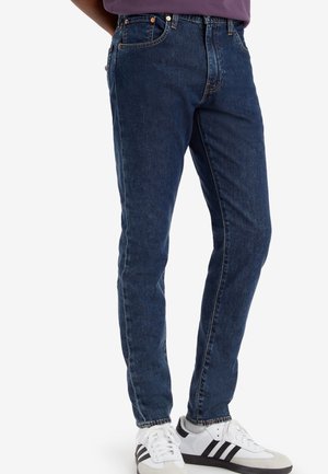Homme portant un jean slim bleu foncé et des baskets blanches avec des bandes noires, debout devant un fond blanc uni.