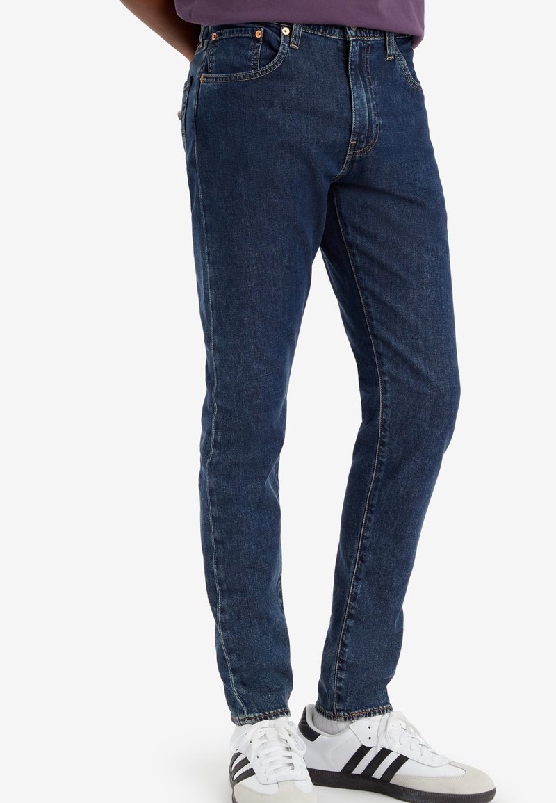 Homme portant un jean slim bleu foncé et des baskets blanches avec des bandes noires, debout devant un fond blanc uni.