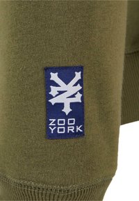 Olijfgroene sweatshirtstof met een gestructureerd oppervlak. Een blauwe patch toont een witte graphic en de tekst "ZOO YORK." Ribgebreide manchetten.