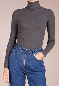 Person, der einen engen, dunkelgrauen, gerippten Rollkragenpullover und hochtaillierte blaue Jeans mit einem Reißverschluss trägt, hat die Hand in der Tasche.