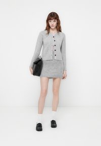 Cardigan grigio a maglia con motivo a trecce, chiusura con bottoni e gonna mini abbinata. Il modello tiene una borsa nera, indossando calze bianche e scarpe nere.