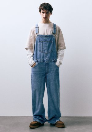 Salopette en jean bleu clair avec bretelles réglables, poche avant et jambes larges. Associée à un t-shirt beige à col rond et des chaussures marron.