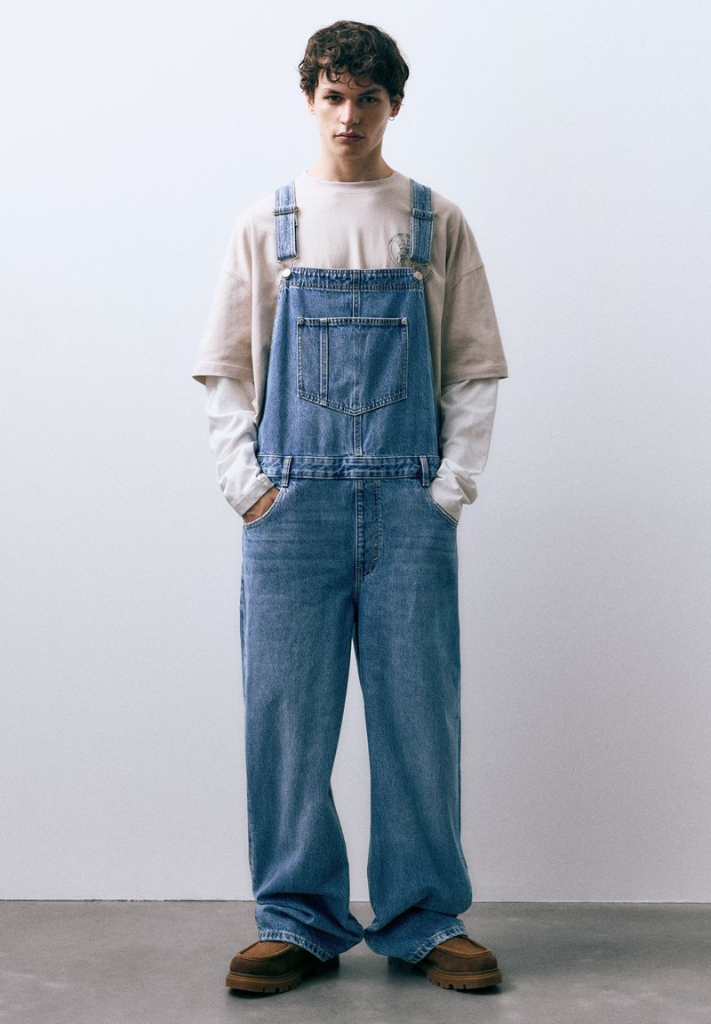 Lichtblauwe spijkeroveralls met verstelbare bandjes, een voorzak en wijde pijpen. Gekleed met een beige shirt met ronde hals en bruine schoenen.