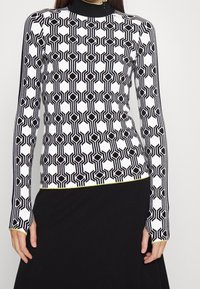 Top de manga larga con un patrón geométrico en blanco y negro con formas hexagonales y un ribete amarillo en el dobladillo. Material liso y elástico.