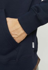 Sweat-shirt marine avec une poche kangourou, des poignets et un ourlet côtelés. Tissu texturé avec une étiquette bien visible sur le côté inférieur.