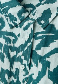 Camicia a maniche corte di un verde menta chiaro con un motivo astratto color te verde, dotata di collo a punta, bottoni bianchi e tessuto testurizzato.