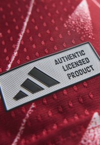 Λευκή ορθογώνια ετικέτα με κείμενο "Authentic Licensed Product" και μαύρο λογότυπο σε υφασμάτινη κόκκινη υφή με μικρές κουκκίδες και λευκές ρίγες.