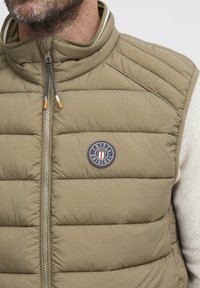 Olivgrüne Pufferweste mit hohem Kragen, ausgestattet mit horizontalen Stepppaneelen und einem runden Logo-Patch. Reißverschluss mit kontrastierenden Zuglaschen.