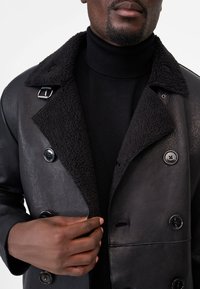 Black leather double-breasted coat with a fuzzy black collar. Features large buttons and a smooth texture. Worn over a black sweater.