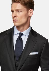 Completo nero su misura con rever a punta, abbinato a una camicia azzurra chiara e a una cravatta blu navy a fantasia. Un fazzoletto bianco nella tasca sinistra aggiunge un tocco di dettaglio.