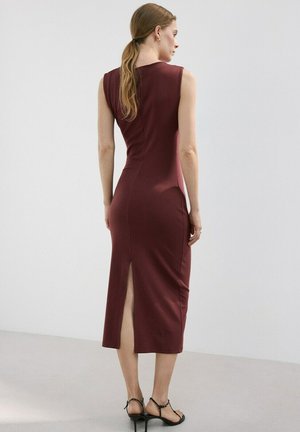 Mujer con cabello largo, vestida con un vestido midi sin mangas color burdeos con una abertura en la parte trasera, calzando sandalias negras de tacón alto, de pie frente a un fondo liso.