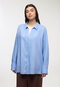 Jasnoniebieska koszula z długim rękawem z gładkiego materiału; ma kołnierzyk i zapięcie na guziki z przodu; noszona na białym topie.