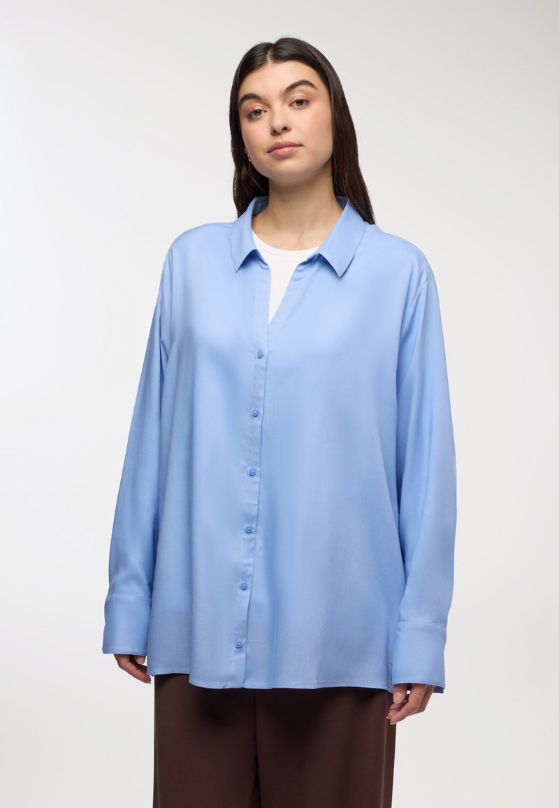 Jasnoniebieska koszula z długim rękawem z gładkiego materiału; ma kołnierzyk i zapięcie na guziki z przodu; noszona na białym topie.
