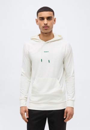 Mann trägt ein weißes Kapuzensweatshirt mit Fronttasche und grünem "30904"-Text, steht vor einem schlichten hellen Hintergrund.