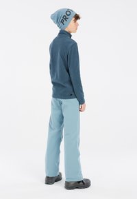 Plavi fleece gornji dio, svijetloplave hlače, sivi čizmi. Beanie s natpisom "PROT" u crnoj boji. Jednostavan dizajn, meka tekstura i praktično za hladno vrijeme.