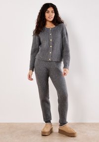 Frau in einem grauen gerippten Strick-Cardigan mit Knöpfen und passenden Hosen, die vor einer schlichten weißen Wand steht und beige Plateauslipper trägt.