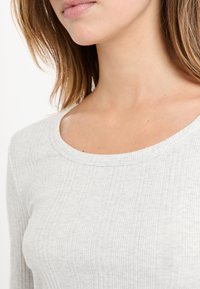 Zbliżenie na szyję, dolną część twarzy i ramię kobiety w jasnoszarym topie z prążkowanej tkaniny z okrągłym dekoltem.