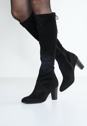 Nogi kobiety w przezroczystych czarnych rajstopach i czarnych zamszowych kozakach na obcasie za kolano z bocznymi zamkami, pozujące na jednolitym białym tle.