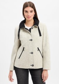 Junge Frau, die eine hellbeige Zip- und Knopfjacke mit schwarzem Kragen und Reißverschlusstaschen trägt und mit neutralem Ausdruck dasteht.