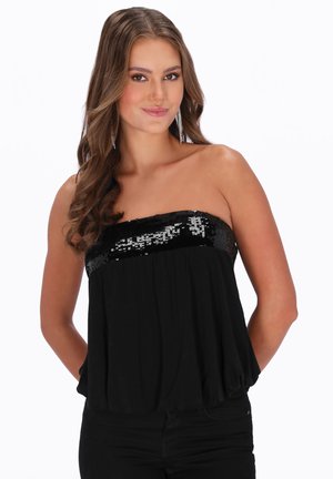 Jonge vrouw met lang golvend haar, gekleed in een zwarte strapless top met pailletten, staand met haar handen achter haar rug tegen een witte achtergrond.