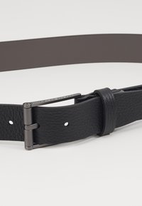 Cinturón de cuero negro con una superficie texturizada, que cuenta con una hebilla y un pasador de metal. El diseño incluye múltiples orificios para ajuste.