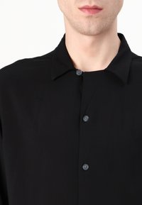 Schwarzes Hemd mit Knöpfen und Kragen, das eine glatte Textur und einen subtilen Glanz aufweist, akzentuiert durch dunkle Knöpfe und ein minimalistisches Design.