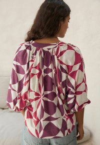 Blouse multicolore en tissu doux avec un motif géométrique de formes rouges et violettes, dotée d'un dos froncé et de manches bouffantes.