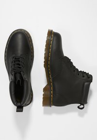 Botas de cuero negro con punta redonda, superficie texturizada y costuras amarillas en contraste. Tienen una suela de goma marrón resistente y un diseño de cordones.